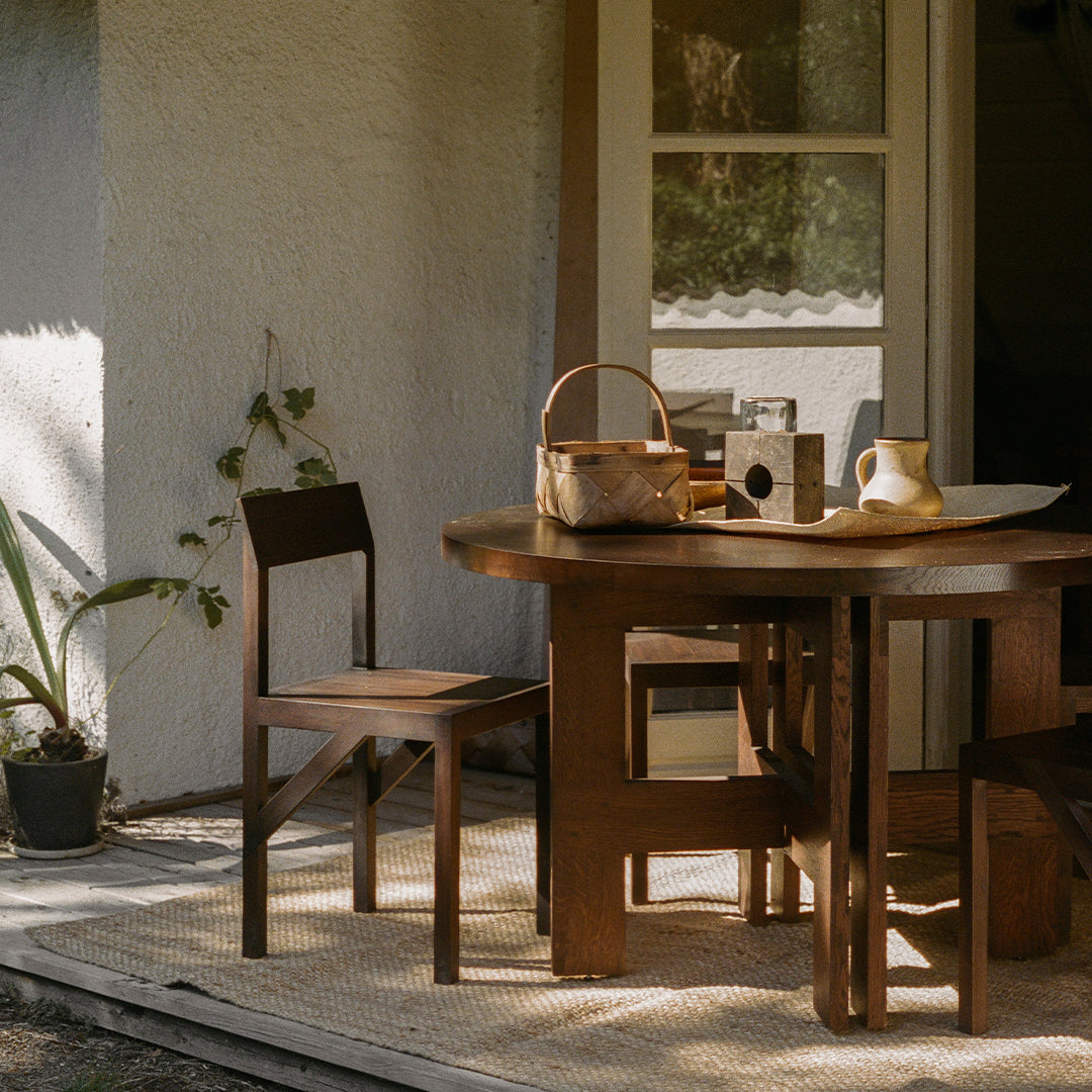 Wooden dining table with rich brown tones, paired with simple chairs, set against a natural outdoor backdrop.