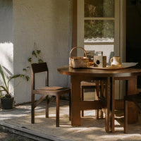 Wooden dining table with rich brown tones, paired with simple chairs, set against a natural outdoor backdrop.