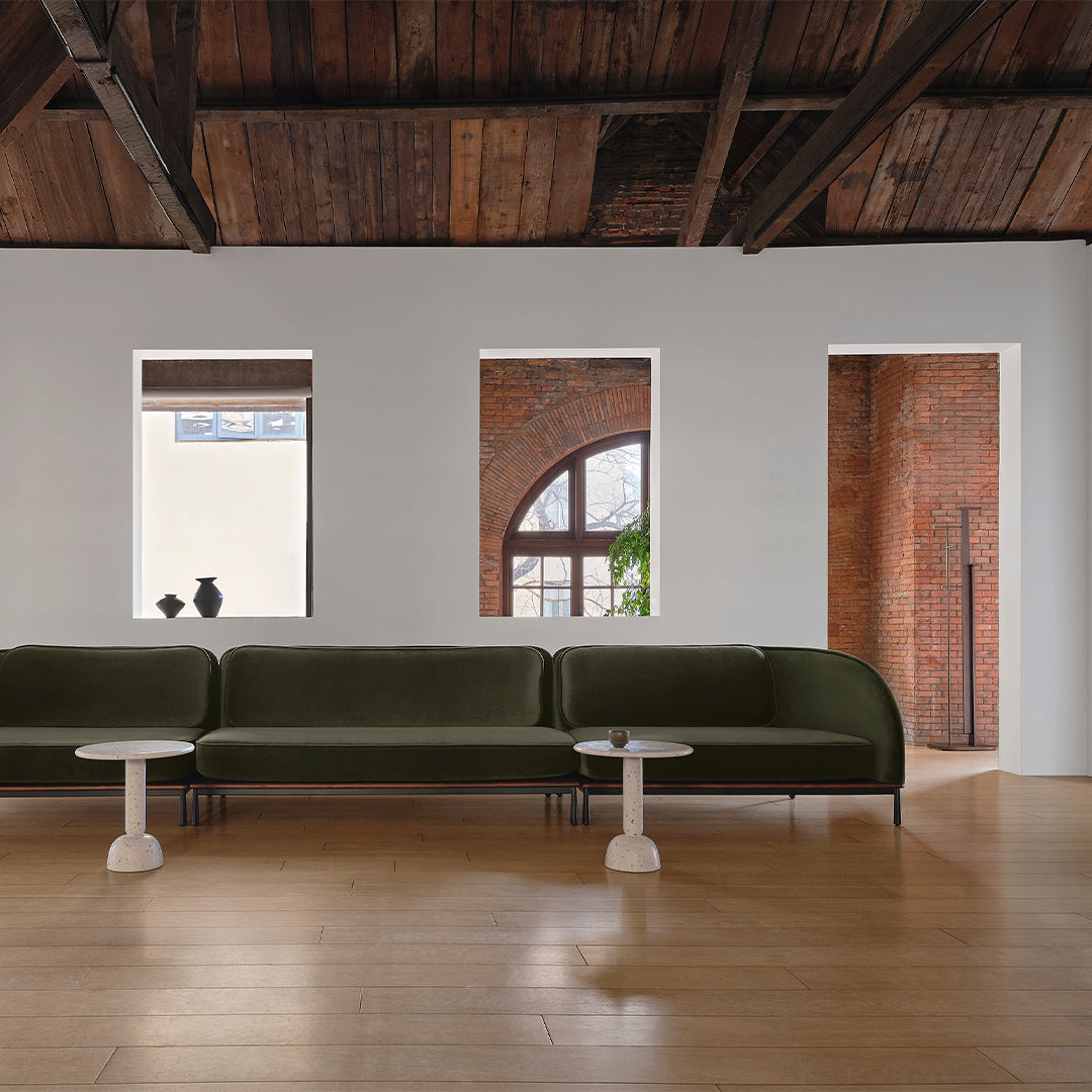 Green velvet sofa with white tables, warm wood flooring, and exposed brick wall accents in a bright room.