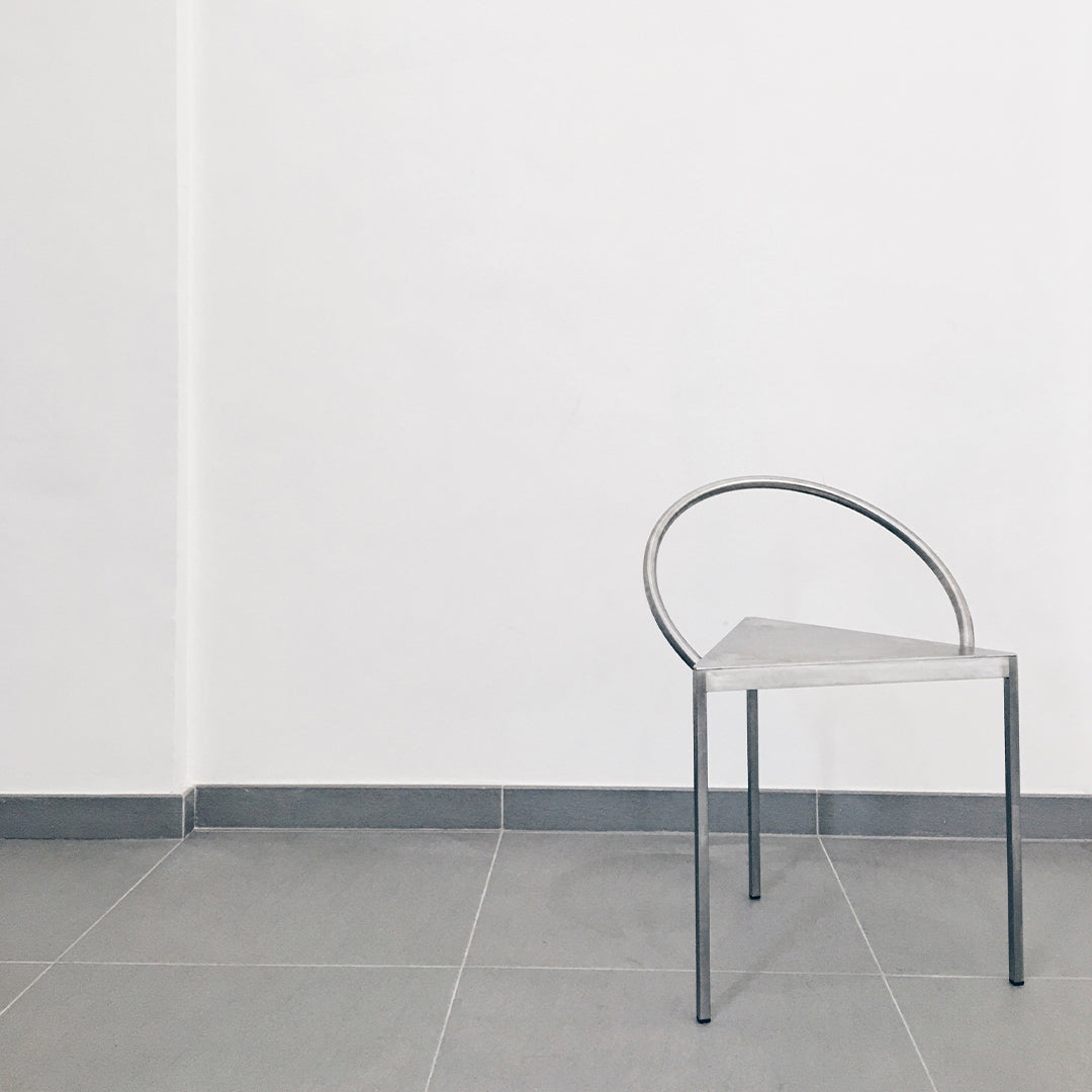 Sleek silver metal chair with a minimalist triangular design, set against a clean white wall and gray tiled floor.