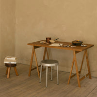 Wooden desk in warm brown tones with a sleek silver stool against a textured beige wall. Minimalist design elements.