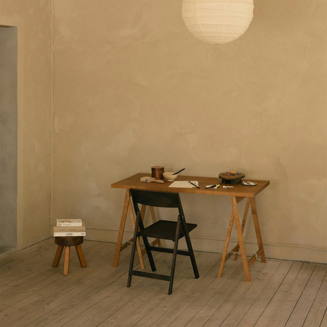 Light beige walls, wooden table in natural finish, black folding chair, and soft white lantern create a neutral, minimalist style.