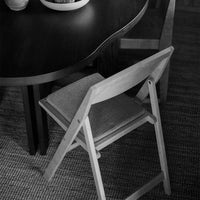 Light wooden folding chair against a dark circular table on a textured rug, creating a contrasting monochrome palette.