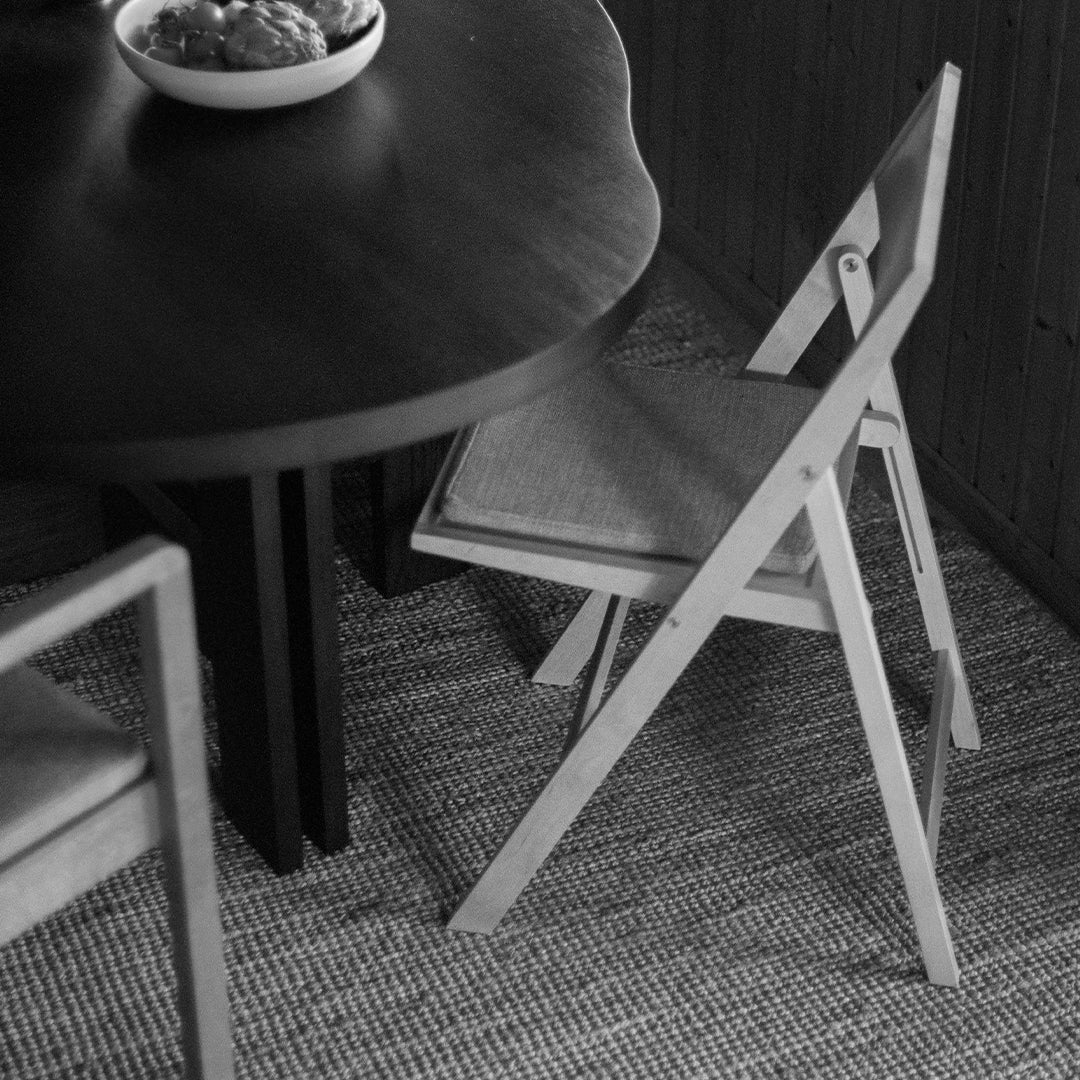 Black and white image featuring a dark wood table, light wood folding chair, and textured woven rug.