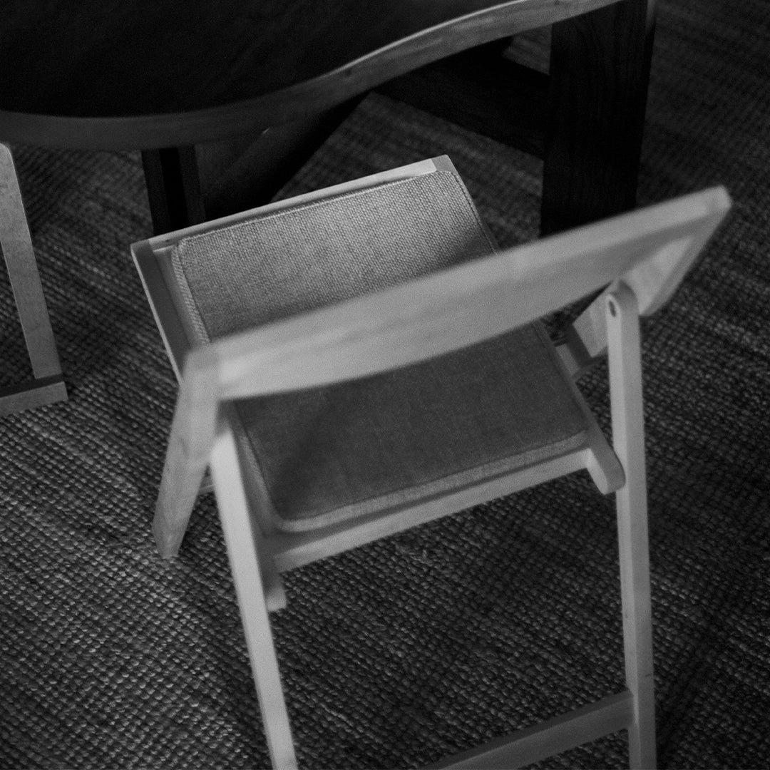 Black and white image of a simple wooden chair with a light gray fabric seat against a textured neutral background.