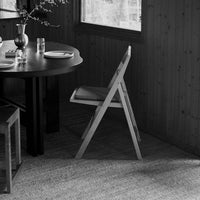 Black and white image of a modern foldable chair with simple lines, placed by a round dark wooden table and textured rug.