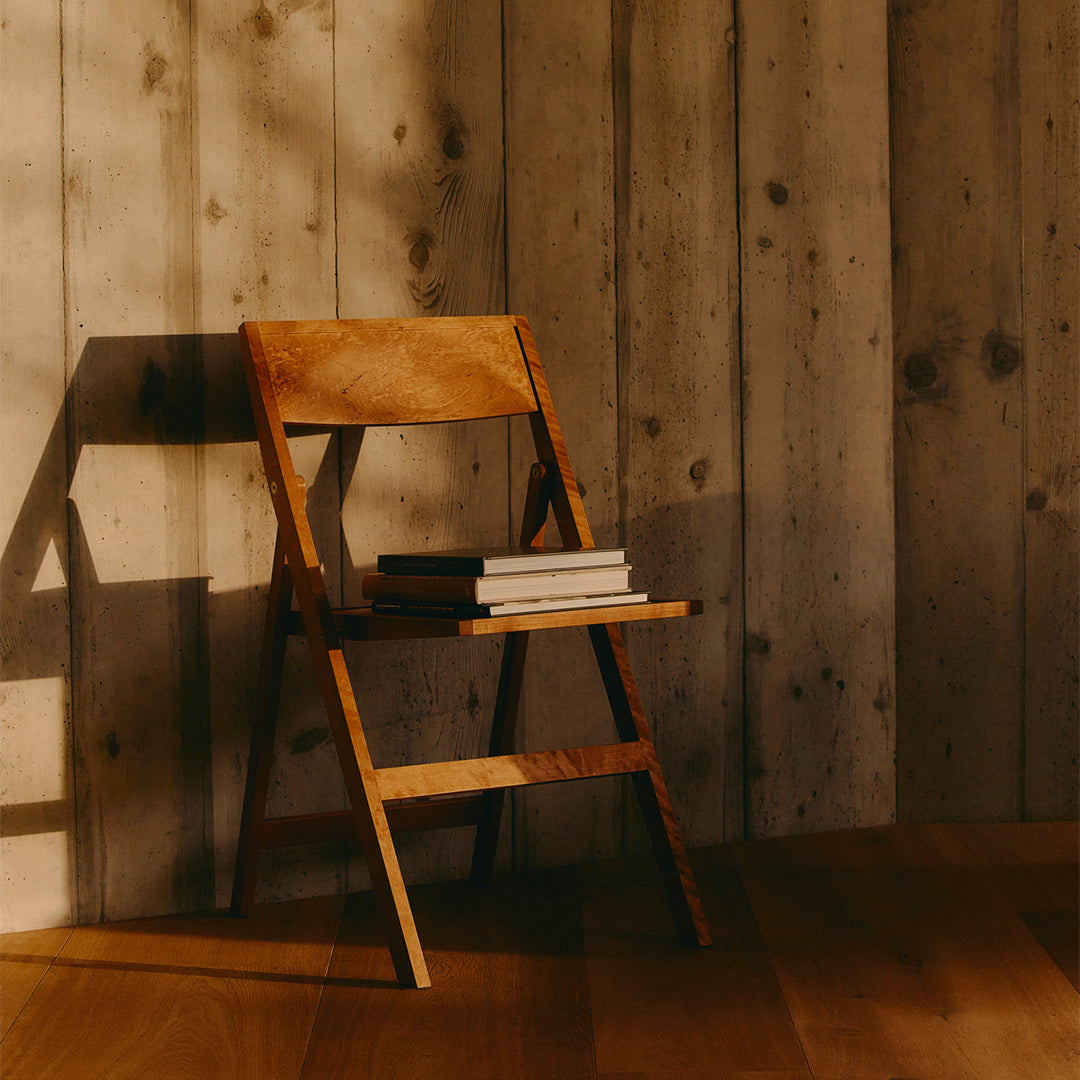 Light-colored wood folding chair against rustic wooden wall with warm, natural tones and soft shadows.