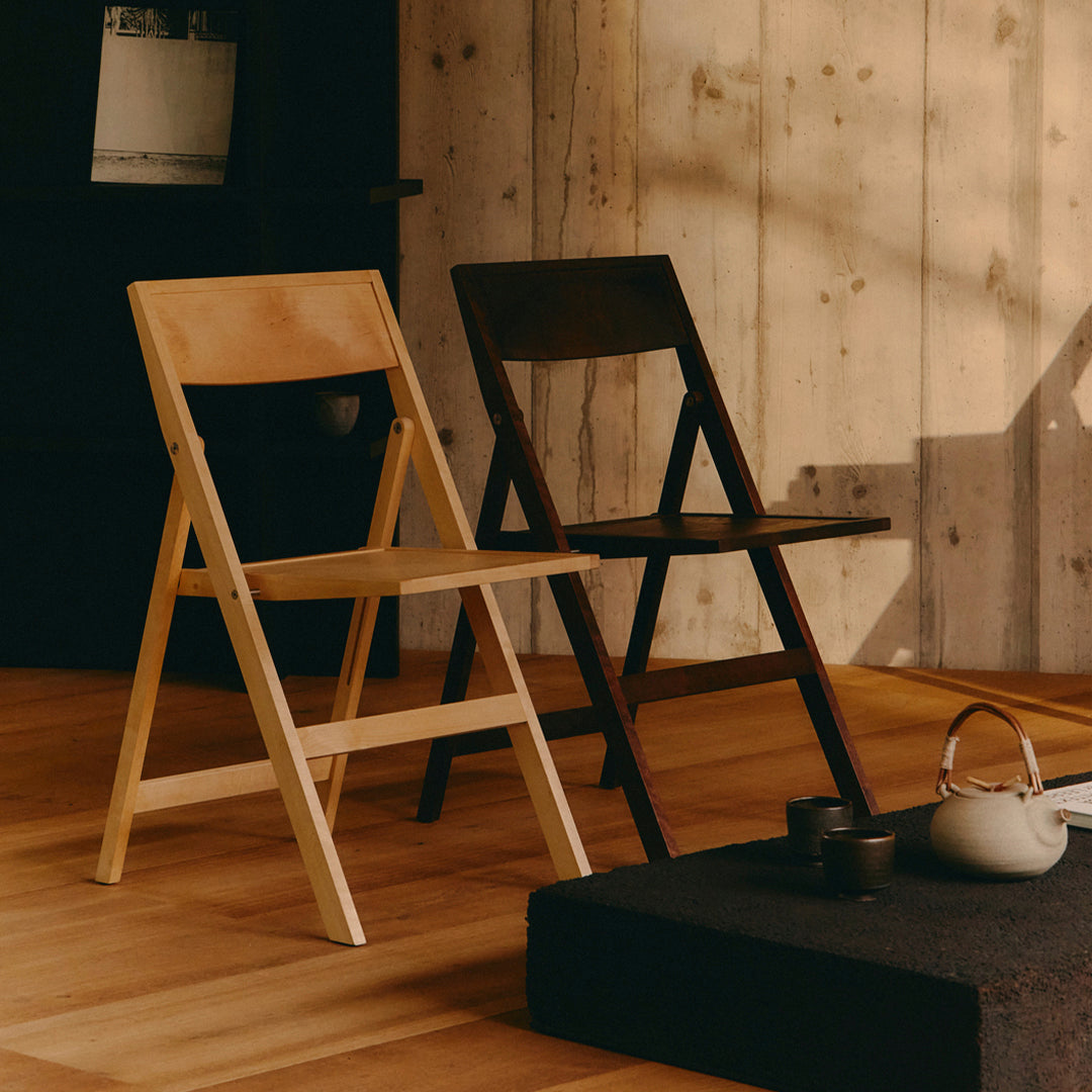 Two stylish folding chairs: one in light natural wood and the other in a dark finish, set against a wood-paneled backdrop.