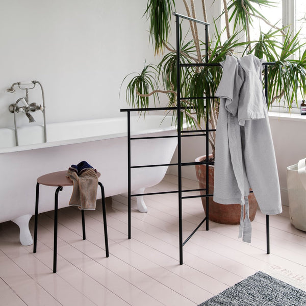 Black clothing rack and stool on light pink floor, surrounded by greenery and a white bathtub for a serene aesthetic.