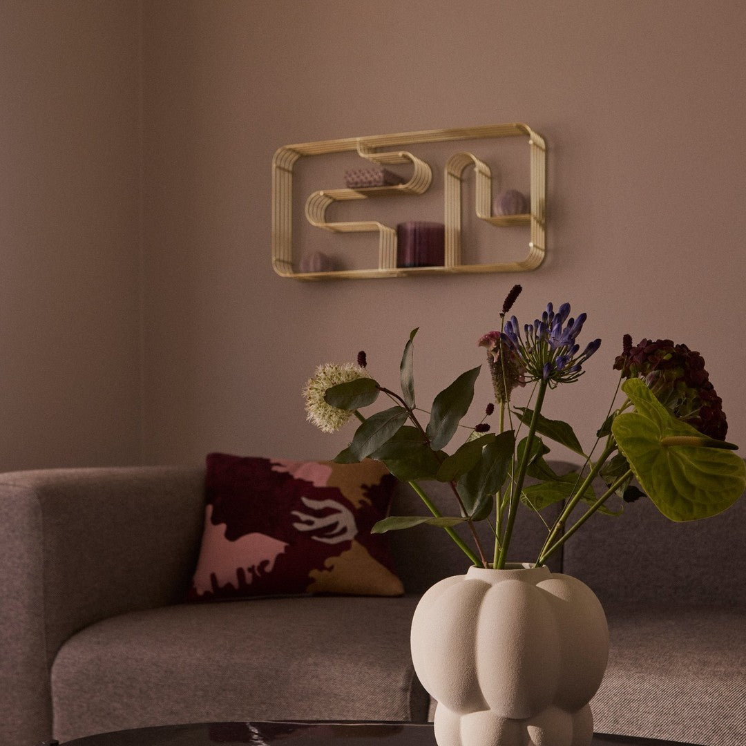 "Beige sofa, burgundy and pink patterned pillow, golden shelf, and pastel floral arrangement against a soft taupe wall."