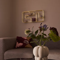 "Beige sofa, burgundy and pink patterned pillow, golden shelf, and pastel floral arrangement against a soft taupe wall."