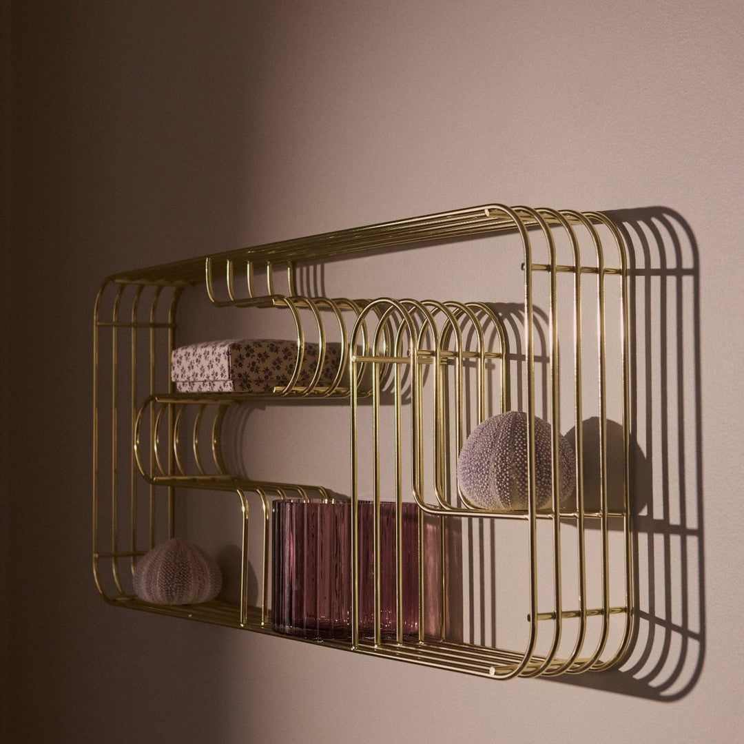 Gold wire shelf with soft pink containers and a floral-patterned box, casting elegant shadows on a wall.