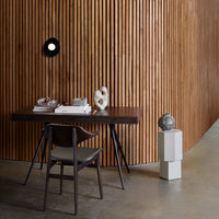 Modern desk with a dark wood finish, complemented by a geometric white pedestal and a textured chair against warm wood paneling.