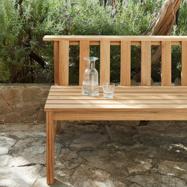 Natural wood bench with a light brown finish, featuring horizontal slats and a rustic stone backdrop.