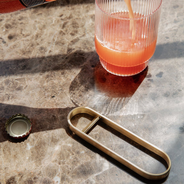 Brass bottle opener on a marbled surface with shadows, featuring a ribbed pink glass filled with juice.