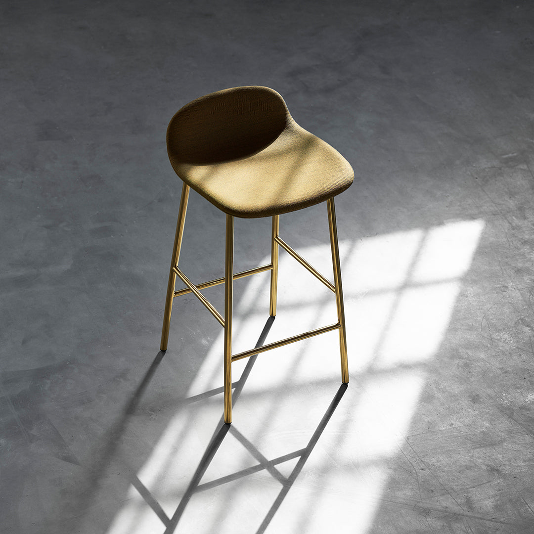 Sleek gold bar stool with a smooth, rounded tan seat and minimalist design, set against a shadowed concrete floor.