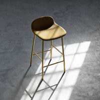 Sleek gold bar stool with a smooth, rounded tan seat and minimalist design, set against a shadowed concrete floor.
