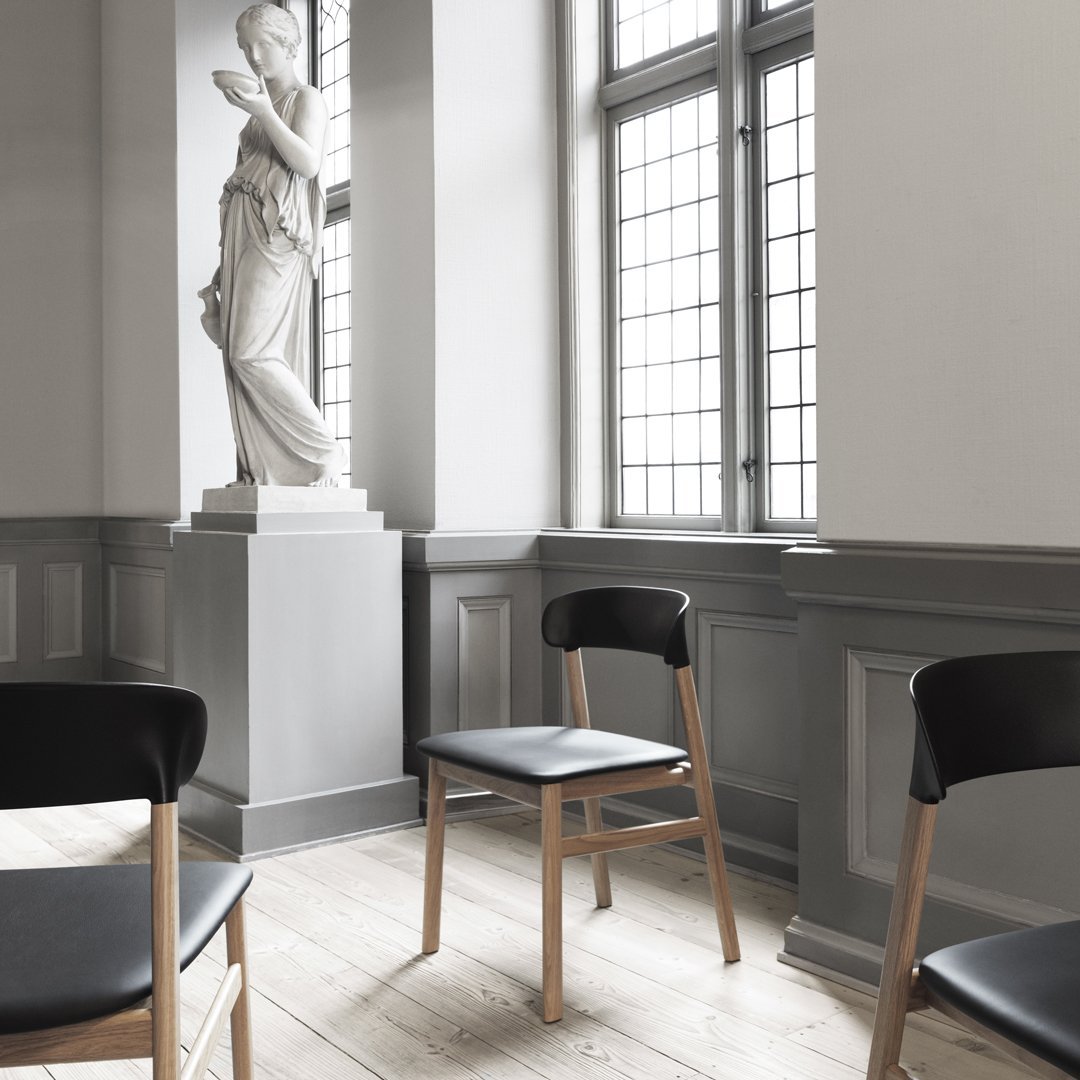Black chairs with wooden legs on light wood flooring, set against grey walls and large windows. Minimalist and modern design.