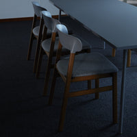 Four gray upholstered chairs with light wood legs beside a dark table on a black carpeted floor.
