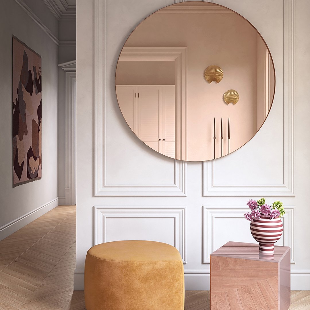 Round rose gold mirror reflecting soft pink walls, paired with a mustard yellow pouf and a geometric patterned vase.