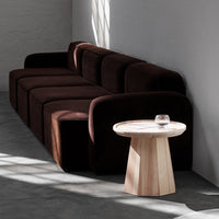Dark brown velvet sofa with soft, rounded shapes, paired with a light wooden side table in a minimalist setting.