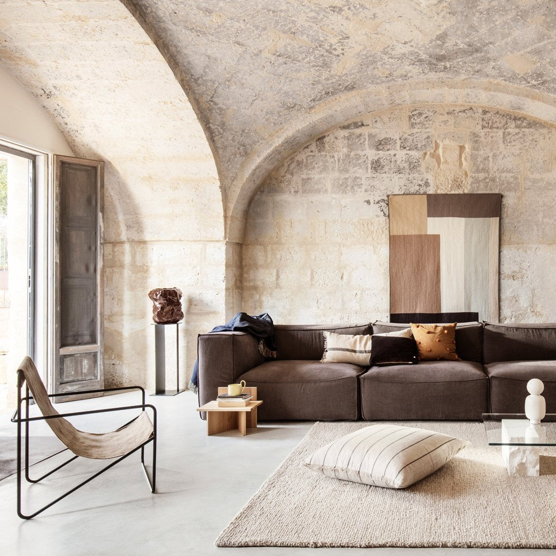 Modern living room featuring brown sofa, neutral patterned rug, and earthy tones in artwork. Natural light enhances warmth.