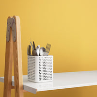 White perforated utensil holder against a vibrant yellow wall, complemented by a sleek white table and wooden easel.