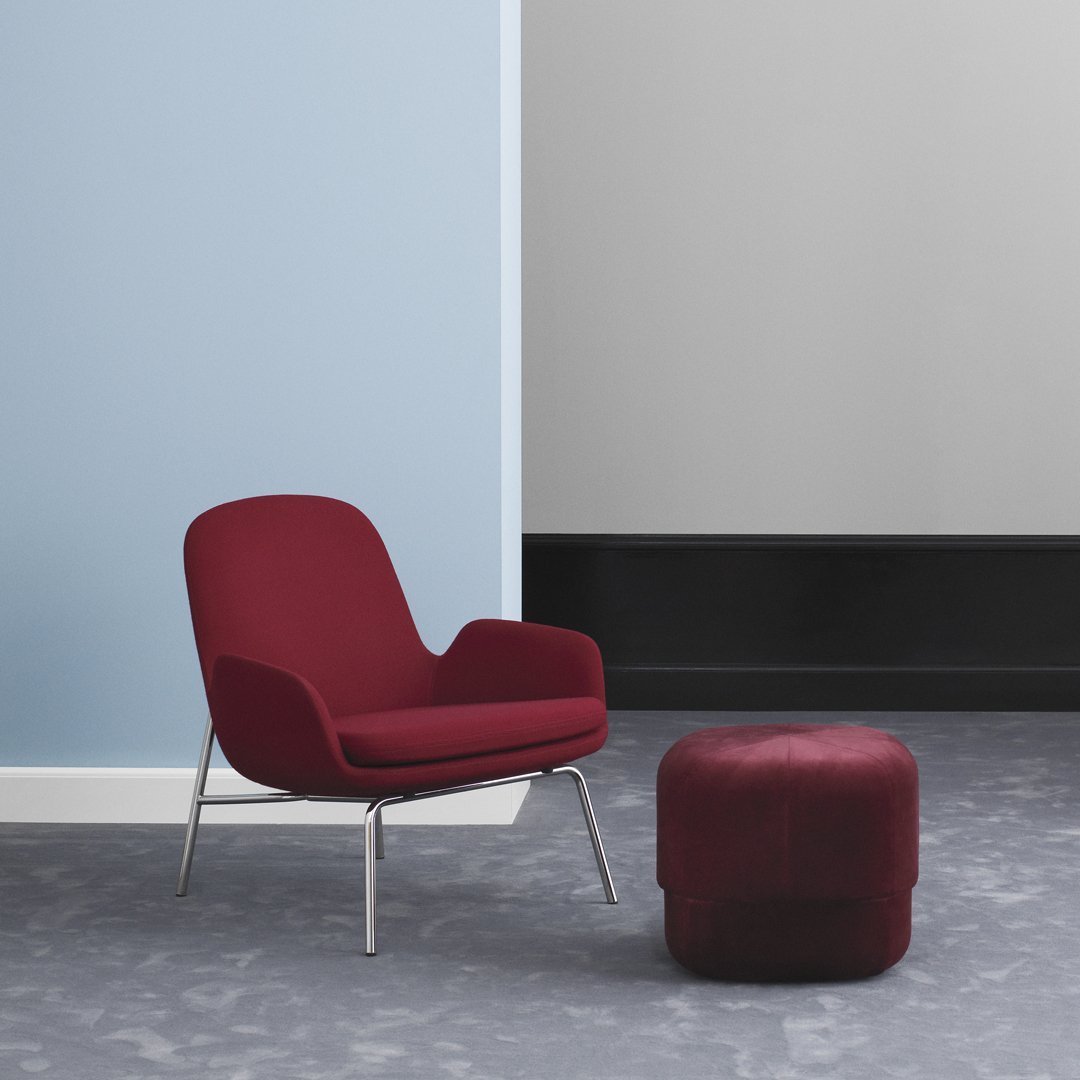 Maroon upholstered chair and pouf on a textured gray floor against a pale blue wall. Modern, minimalist design.