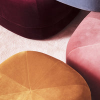 Colorful velvet ottomans in rich burgundy, soft pink, and warm mustard geometric patterns, set on a light textured surface.
