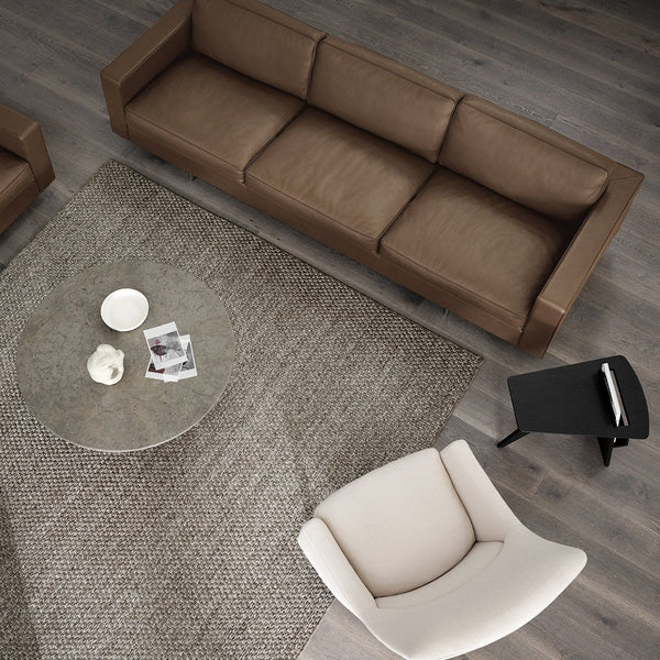 Brown modern sofa, beige accent chair, clear glass coffee table, and textured gray area rug on wood floor.
