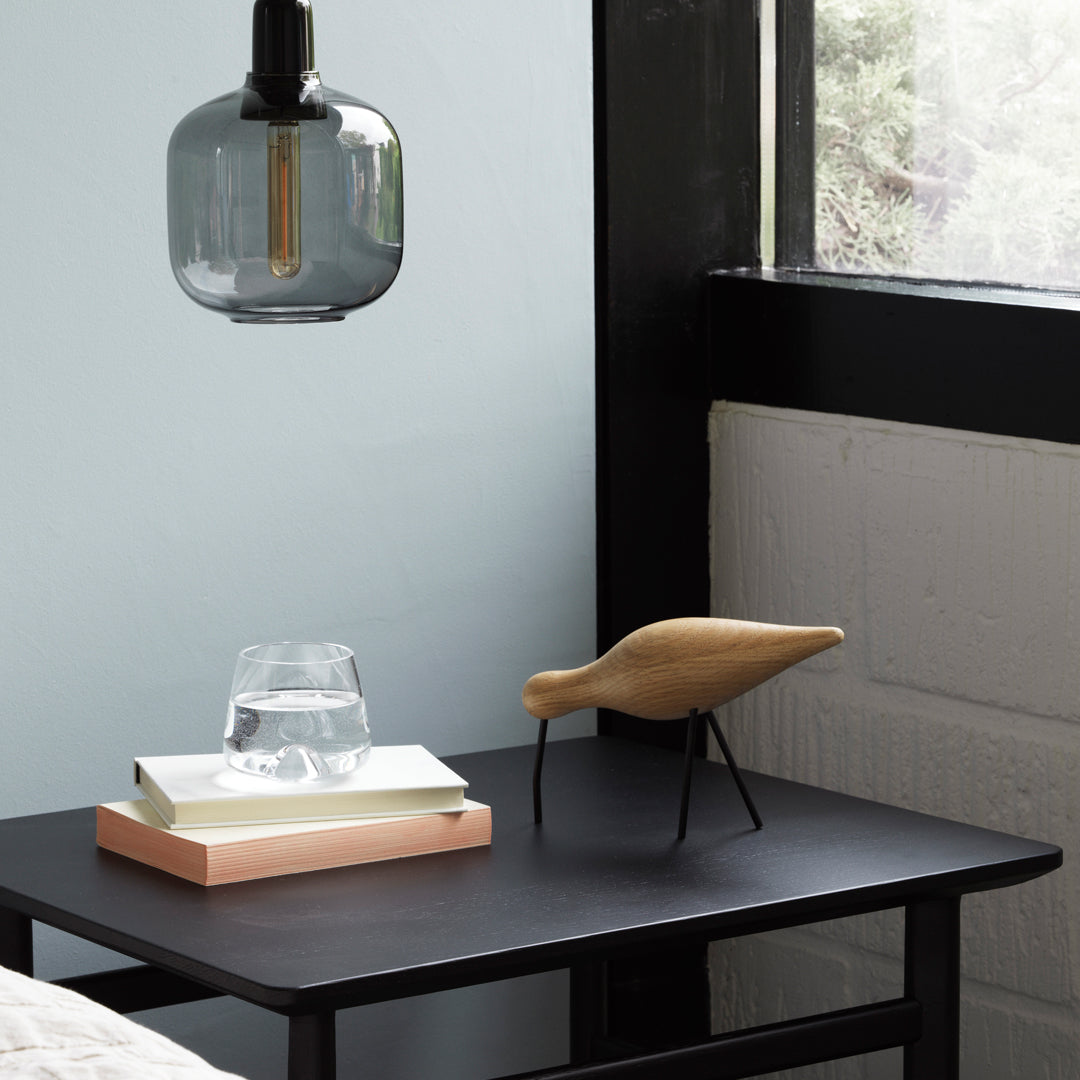 Wooden bird sculpture on black table with a clear glass, minimalist lamp, and pastel background. Modern decor.