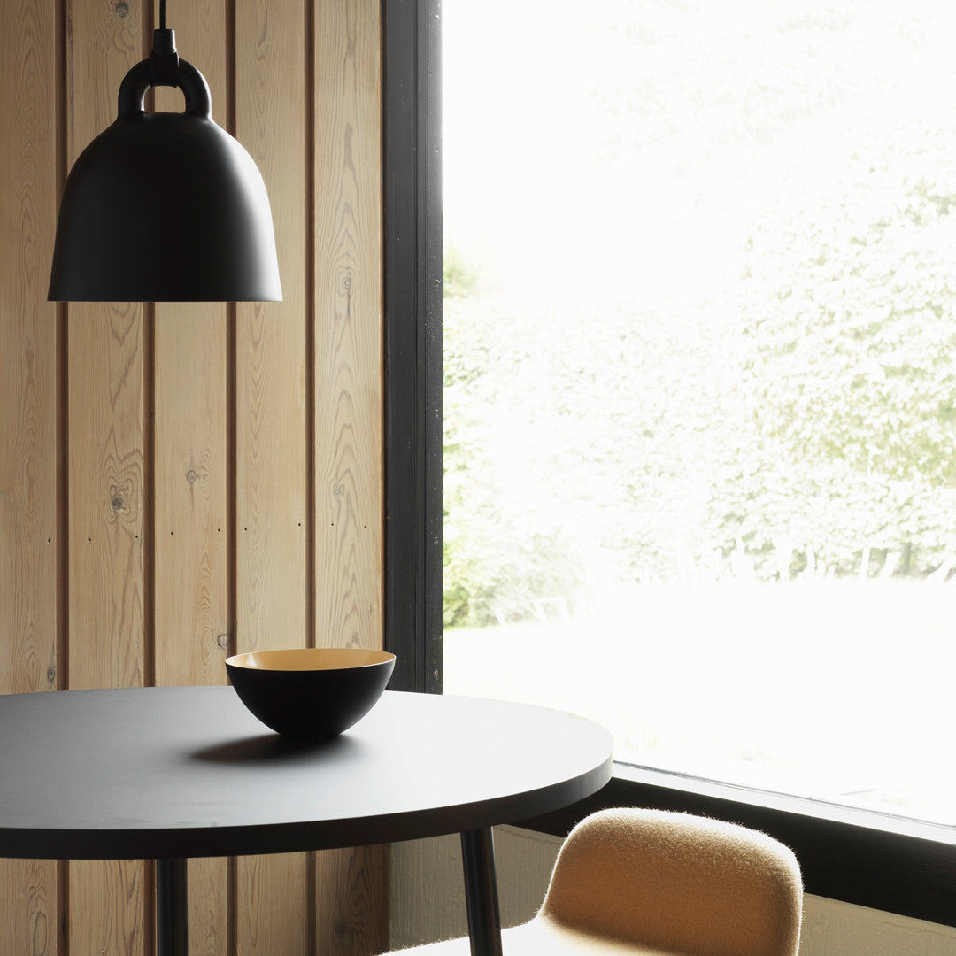 Black pendant light, round black table, and a yellow-accented bowl on a natural wood backdrop with soft natural light.