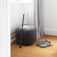 Gray plush ottoman with a black accent, paired with sleek gray shoes on warm beige carpet under soft gray curtains.