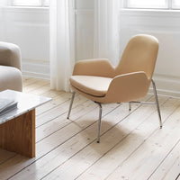 Beige lounge chair with smooth, curved lines and polished chrome legs, set against light wood flooring and soft drapes.