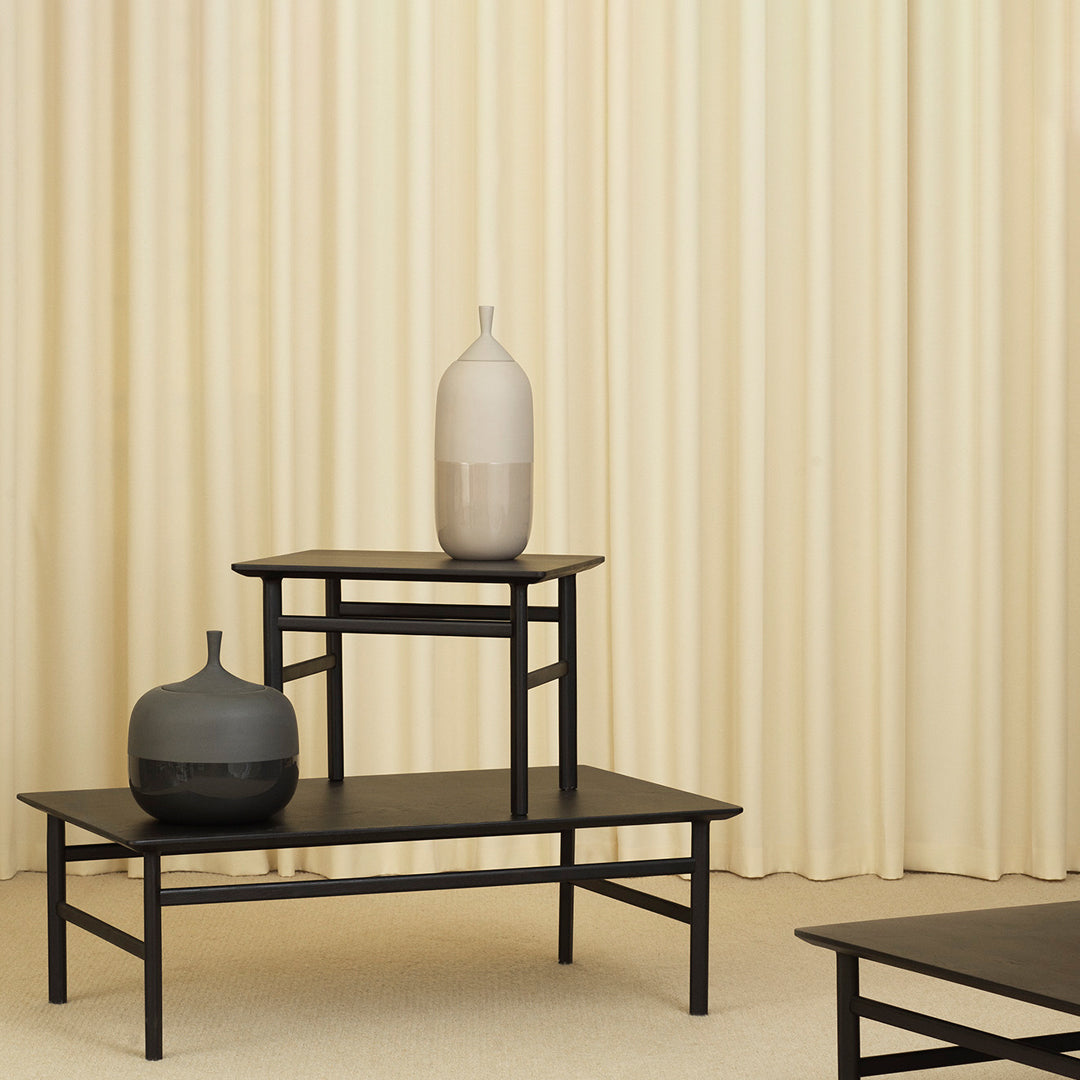 Black and gray vases on black tables with a soft cream backdrop and textured vertical curtain pattern.