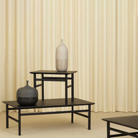 Black and gray vases on black tables with a soft cream backdrop and textured vertical curtain pattern.