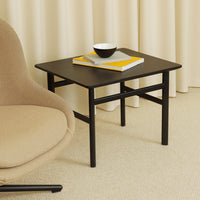 Black square side table with minimalist design, paired with a white bowl and yellow accents, set against neutral tones.
