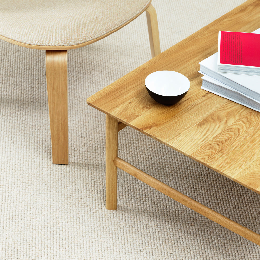 Natural wood coffee table with light beige chair and textured rug; minimalist design with warm oak tones and simple patterns.
