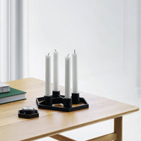 Black geometric candle holder with white candles on a wooden table, complemented by green books and a minimalist setting.