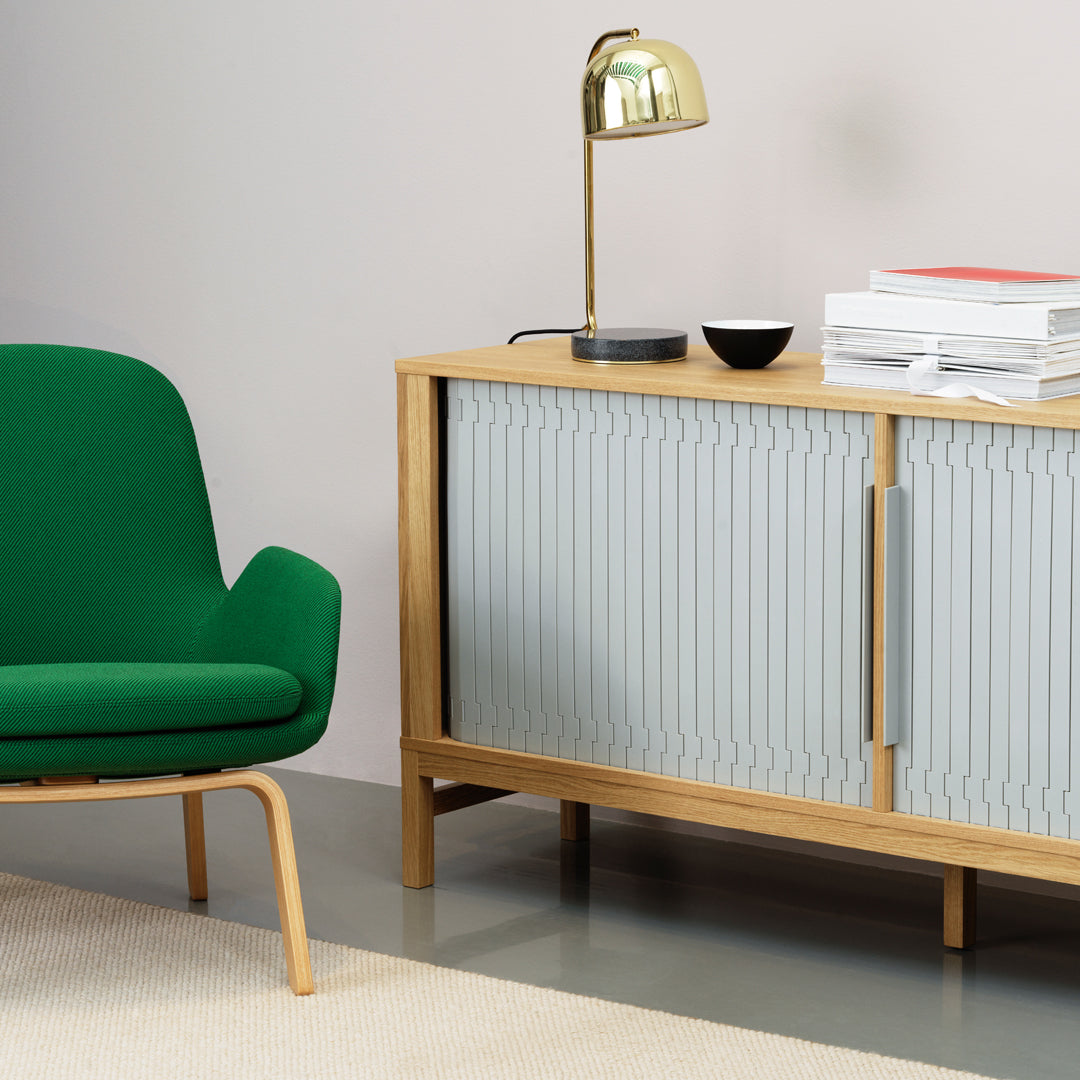 Green chair with curved design, light wood cabinet with ribbed grey doors, gold lamp, minimalist decor.