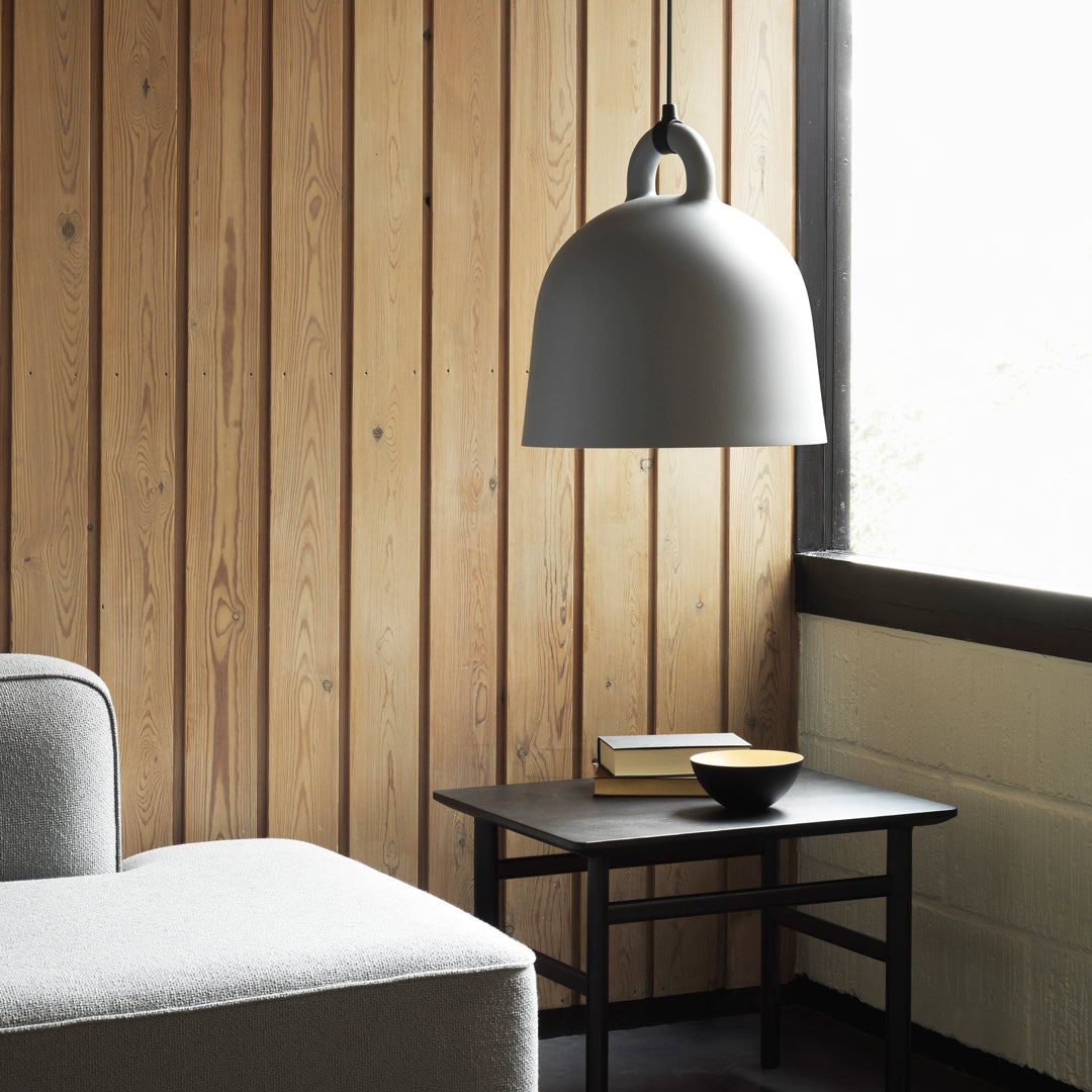 Gray pendant lamp hanging above a black table with a yellow book and bowl, set against light wood paneling.