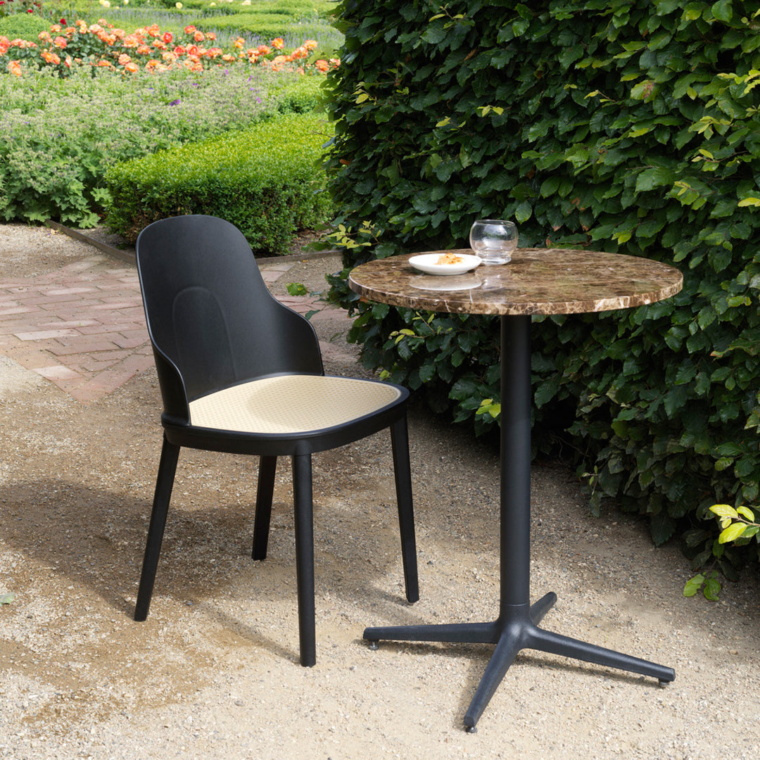 Black chair with cream cushion beside a brown marble tabletop on a sleek black base, set against lush greenery.