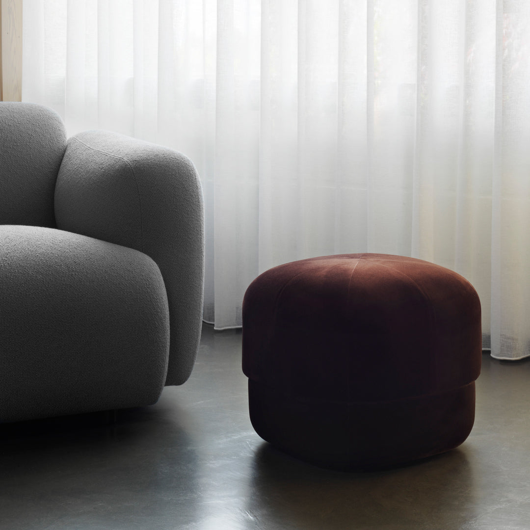 Brown velvet pouf beside a light gray armchair, showcasing a soft, textured finish in a minimalistic interior.