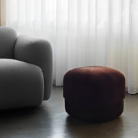 Brown velvet pouf beside a light gray armchair, showcasing a soft, textured finish in a minimalistic interior.