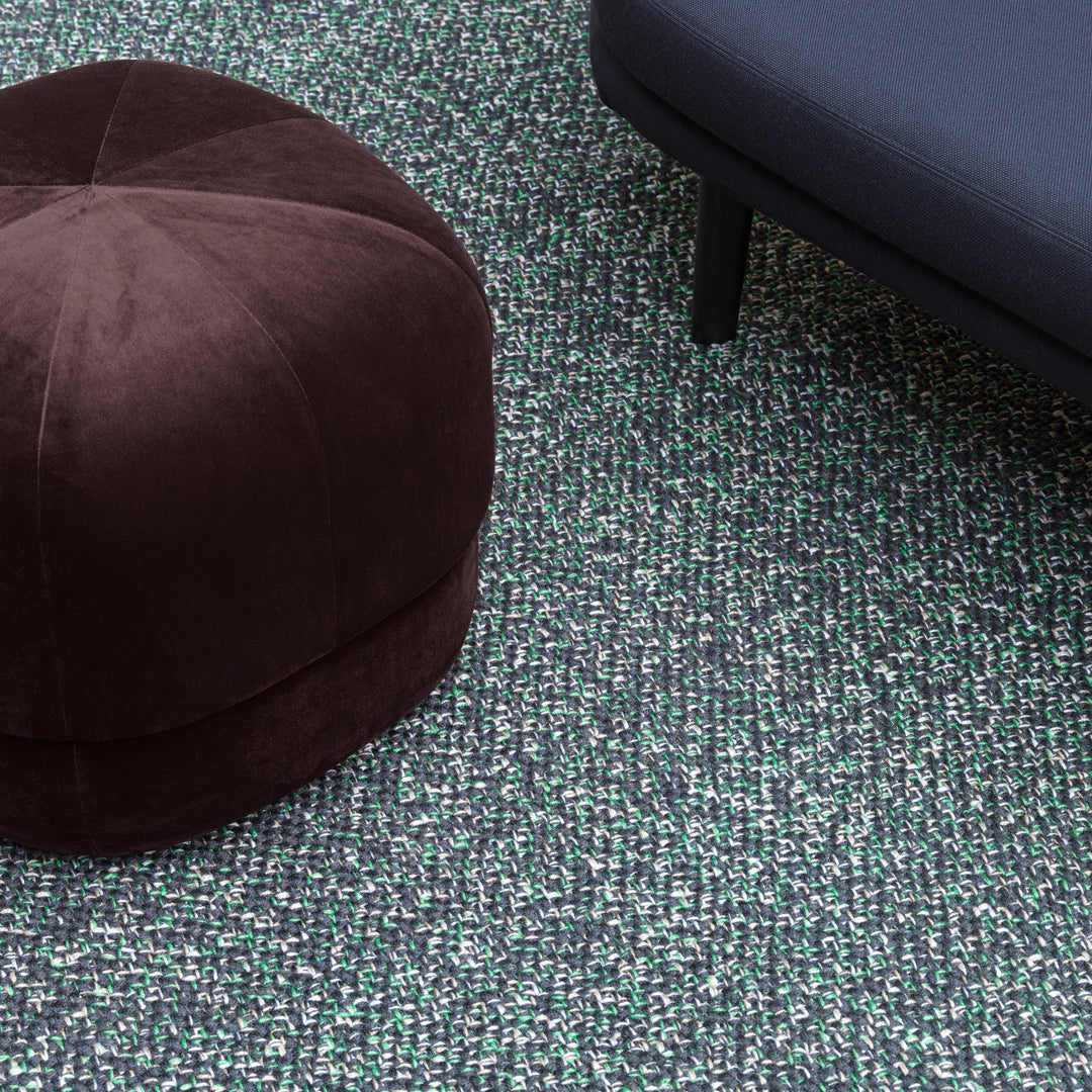 Brown velvet pouf on textured green and black patterned rug with sleek black sofa.