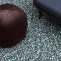 Brown velvet pouf on textured green and black patterned rug with sleek black sofa.