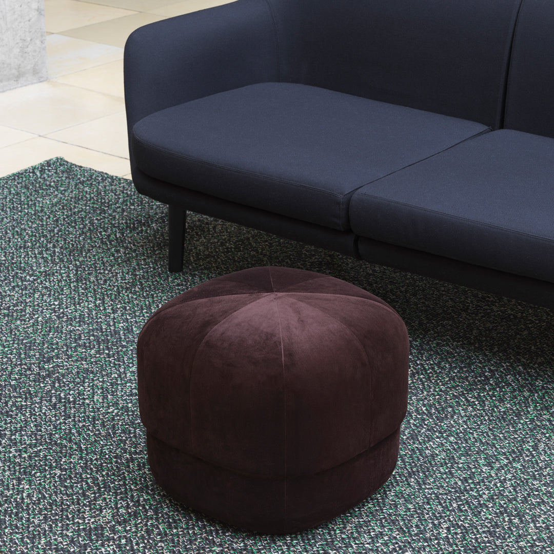 "Round brown velvet pouf on a textured green rug with a dark blue sofa in the background."