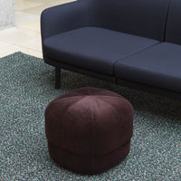 "Round brown velvet pouf on a textured green rug with a dark blue sofa in the background."