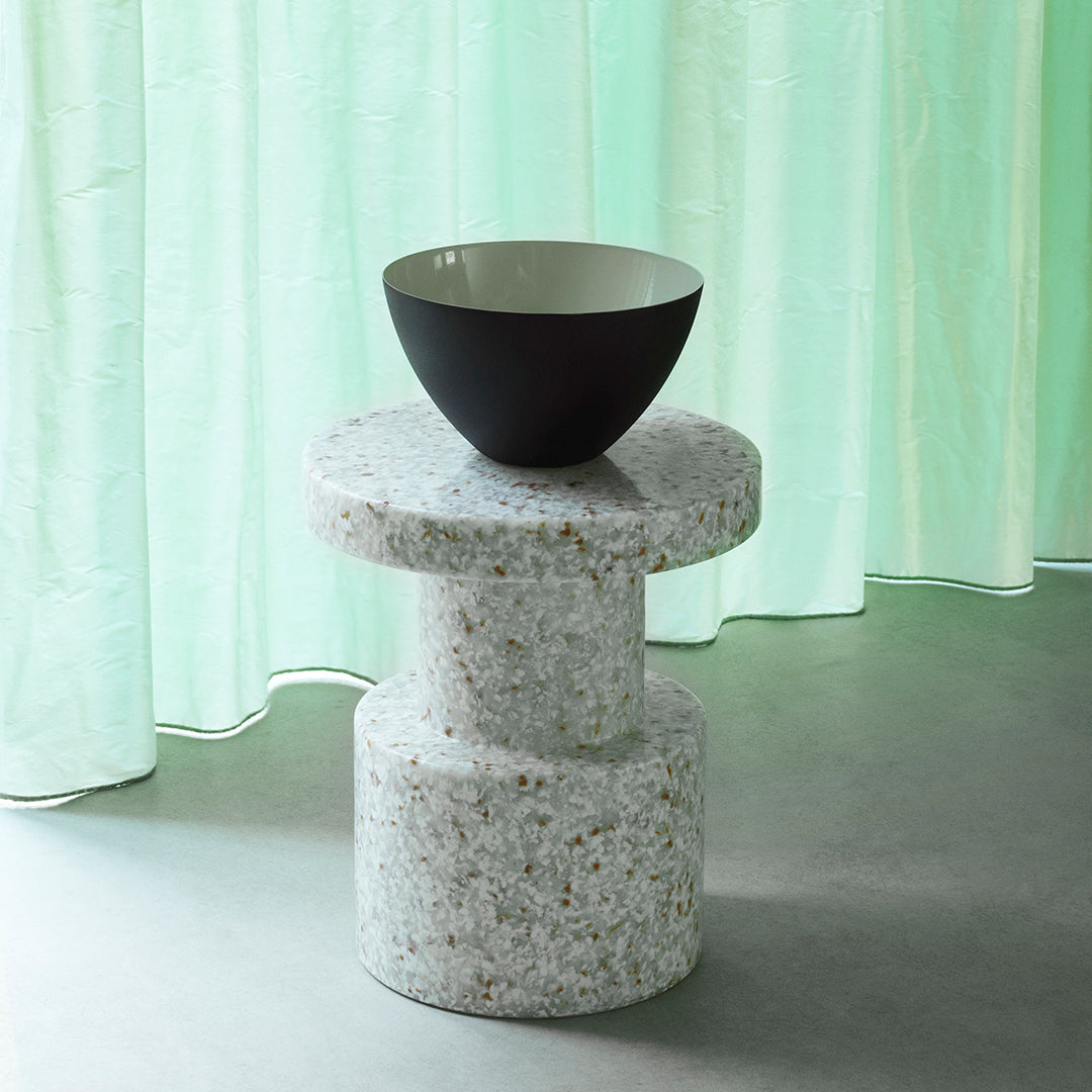 "Round terrazzo side table in white with colorful speckles, topped with a black bowl against soft green curtains."