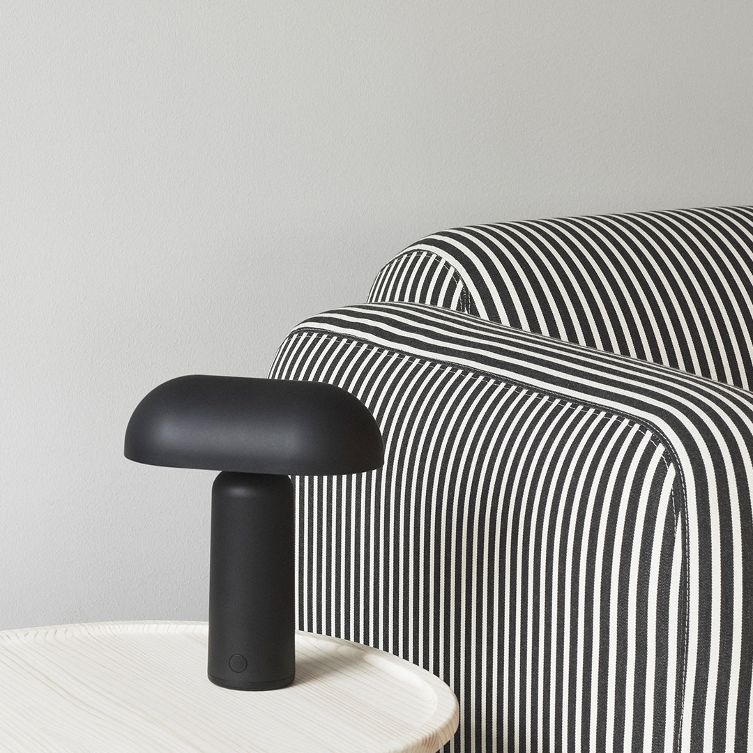 Black mushroom lamp on a white table beside a black and white striped sofa in a minimalist setting.